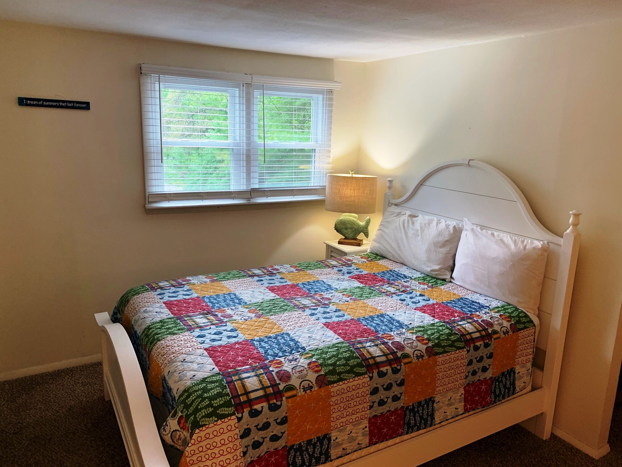 Ein Schlafzimmer mit einem weißen Bett, einer farbenfrohen Steppdecke, einem Nachttisch mit Lampe und einem Fenster mit Jalousien.