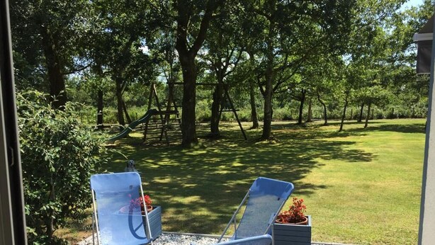Une terrasse avec deux chaises bleues et une petite table, donnant sur un espace verdoyant agrémenté d’arbres et d’une aire de jeux.