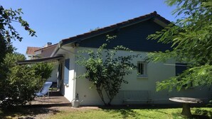 Une maison de plain-pied avec un toit sombre, un petit porche et un jardin agrémenté d’un banc et d’une table.