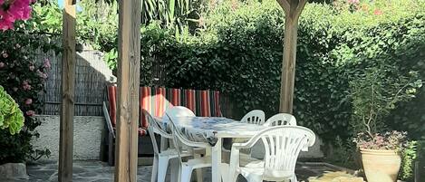 A stone patio with a pergola, white plastic chairs, and a table.