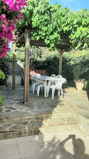 Eine Steinterrasse mit einer Pergola, weißen Kunststoffstühlen und einem Tisch.