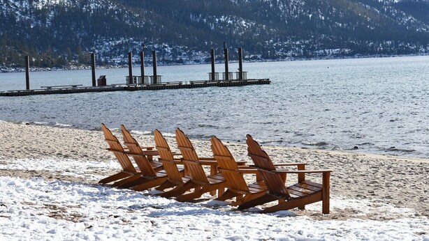 En strand med trästolar och en brygga som sträcker sig ut i en sjö, omgiven av snötäckta berg.