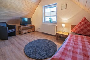 A cozy room with a television on a wooden stand, a window with blinds, a radiator, a lamp on a bedside table, and a checkered bedspread.