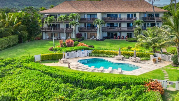 A multi-story hotel with a swimming pool, surrounded by lush greenery and palm trees. - Spacious 2BR Condo | Oceanfront Lanai | Washer/Dryer | BBQ (Kapaa)