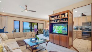 A spacious living room with a large sectional sofa, a coffee table, and a flat-screen TV. There is a wooden entertainment center with framed pictures and a television displaying a tropical scene. The room has a ceiling fan, large windows with blinds, and a view of the ocean. - Spacious 2BR Condo | Oceanfront Lanai | Washer/Dryer | BBQ (Kapaa)
