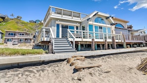 A beachfront house with a balcony, large windows, and a staircase leading to the entrance. - Comfortable Beach House on the Sand (Aptos)