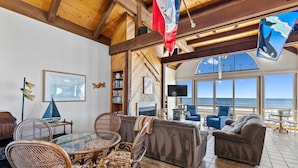 A spacious living area with a wooden ceiling, a glass table, wicker chairs, a brown sofa, and a view of the ocean through large windows. - Comfortable Beach House on the Sand (Aptos)