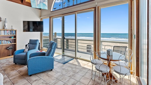 A beachside room with a large sliding glass door to the ocean, a TV, a bookshelf, and a dining area with metal chairs. - Comfortable Beach House on the Sand (Aptos)