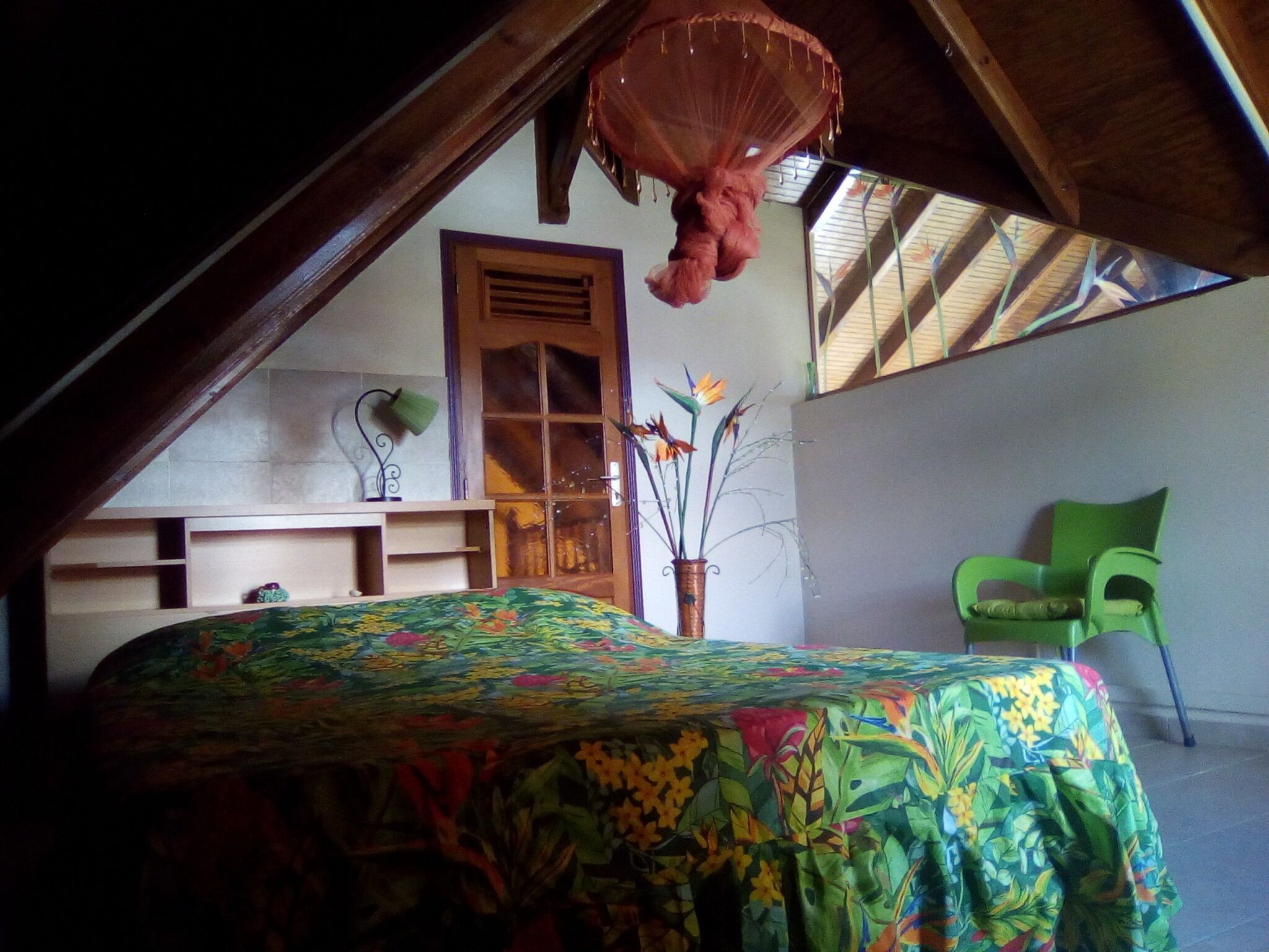 Une chambre avec un plafond en pente, un lit recouvert d’une couvre-lit à motifs floraux, une chaise verte et une porte en bois.