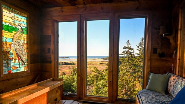 Una cabaña con un amplio ventanal que ofrece vistas a un bosque y a la playa. En su interior, hay un banco de madera, una mesa con un cuenco de conchas marinas y un sofá estampado.