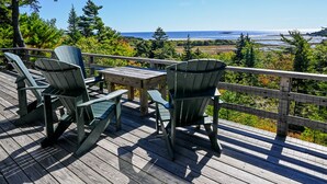 Una terraza de madera con dos sillas Adirondack y una mesa, con vista a un paisaje costero con árboles y el océano.