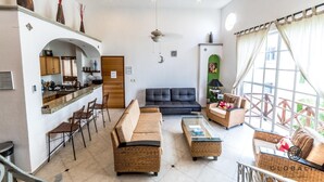 Un vaste espace de vie comprenant un canapé, deux fauteuils en osier, une table basse et un coin repas avec un comptoir et des chaises.
