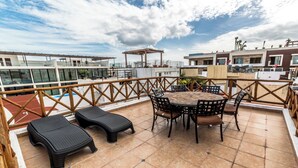 A rooftop terrace with a round table and chairs, a couple of lounge chairs, and a wooden railing.