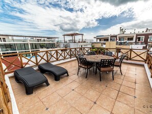 A rooftop terrace with a round table and chairs, a couple of lounge chairs, and a wooden railing.