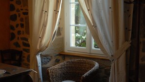 Une pièce aux murs de pierre, avec un fauteuil en osier, une table et une fenêtre ornée de rideaux.