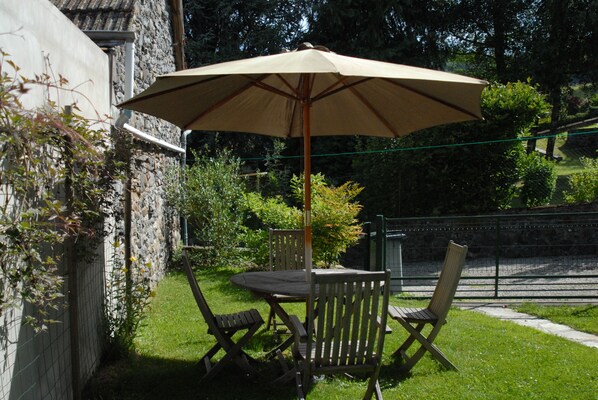 A patio with a table and chairs under a large umbrella, surrounded by greenery and a stone wall. - House 1850 renovated in 2003 located in the village centre (Saignes)