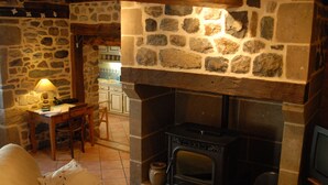 Une cheminée en pierre avec un manteau en bois, une table à manger avec des chaises et une lampe.
