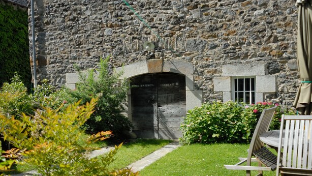 Un bâtiment en pierre doté d’un portail central en arc de cercle, entouré d’un jardin bien entretenu et agrémenté de mobilier d’extérieur.