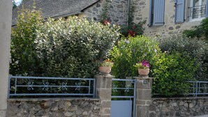 Un mur de pierre surmonté d’un portail en métal, un bâtiment en pierre avec des volets bleus et une végétation luxuriante.