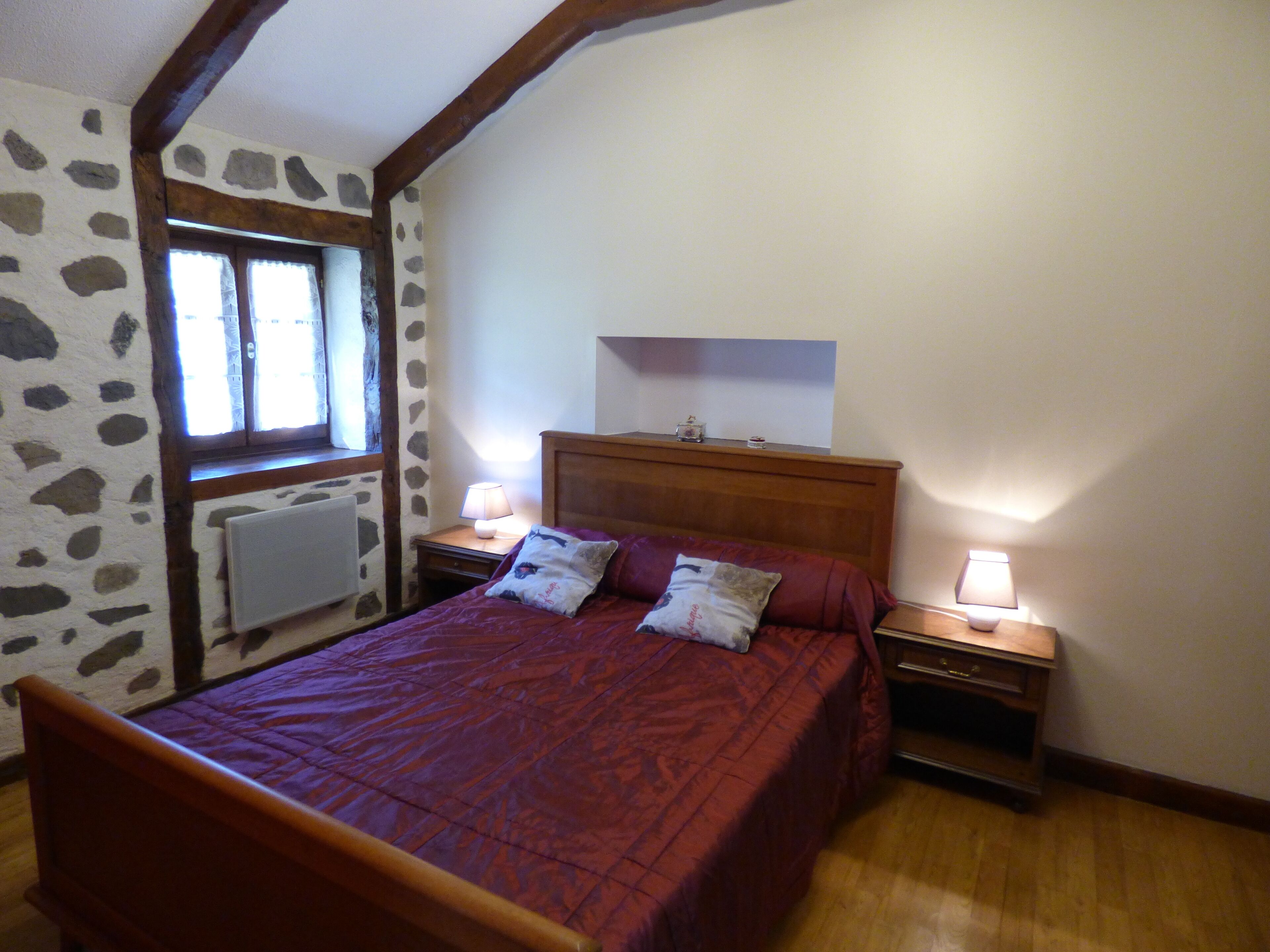 Un dormitorio con una cama de madera, dos mesitas de noche con lámparas, una pared de piedra, una ventana y una viga de madera en el techo.