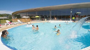 Piscina com espreguiçadeiras e guarda-sóis, cercada por um edifício moderno com grandes janelas.