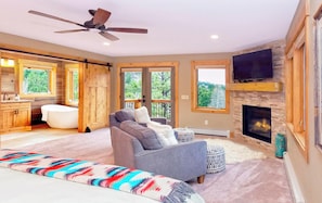 A spacious bedroom with a large bed, a gray sofa, a fireplace, and a TV mounted on the wall. - Luxury House, River Access, RMNP, Mountain Views, Hot Tub! Dancing Pines, Estes Park CO (Estes Park)