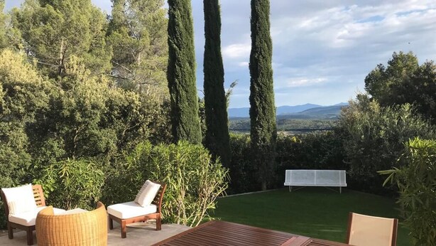 En udendørs terrasse med et træbord og stole, omgivet af grøn vegetation og udsigt til fjerne bjerge.