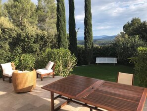 An outdoor patio with a wooden table and chairs, surrounded by greenery and a view of distant mountains. - Charming Villa with Spa and Panorama (Lorgues)
