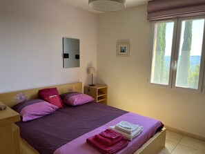 Une chambre à coucher avec un lit, des oreillers, une table de chevet, une lampe, une petite étagère, un cadre photo et une fenêtre donnant sur un beau paysage.