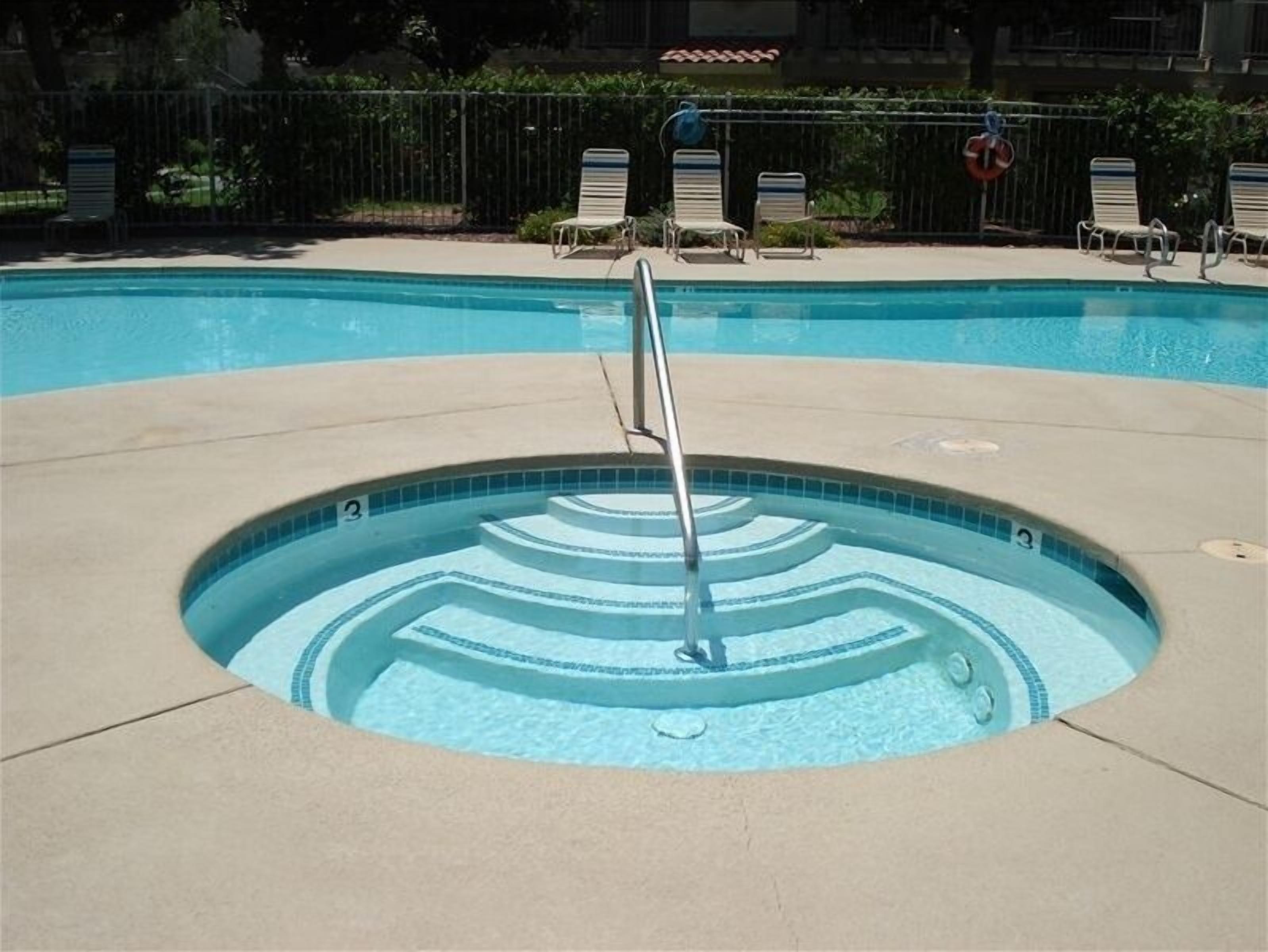 Ein Schwimmbecken mit einer zentralen Wasserspende, umgeben von Liegestühlen und einem Zaun.