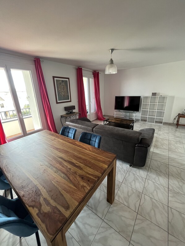 A spacious living room with a wooden dining table, grey sofa, and a TV on a stand.
