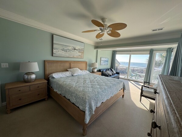 Un dormitorio con cama, mesitas de noche, lámpara, ventilador de techo y vista al océano a través de amplios ventanales.