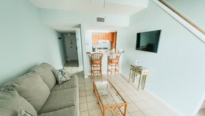 Un salon comprenant un canapé, une table basse et des fauteuils. Il y a également un coin cuisine avec une cuisinière, un évier et des placards. Un téléviseur est fixé au mur.