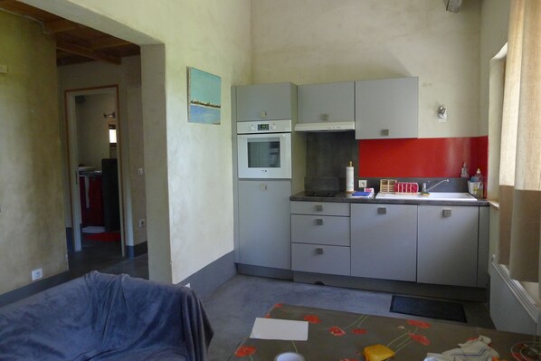 A compact kitchen with a built-in oven, a microwave, and a red backsplash. There is a dining table with a floral tablecloth and a few items on it. - Charming Mazet, completely renovated, very quiet, in pine forest, near the center (Nîmes)