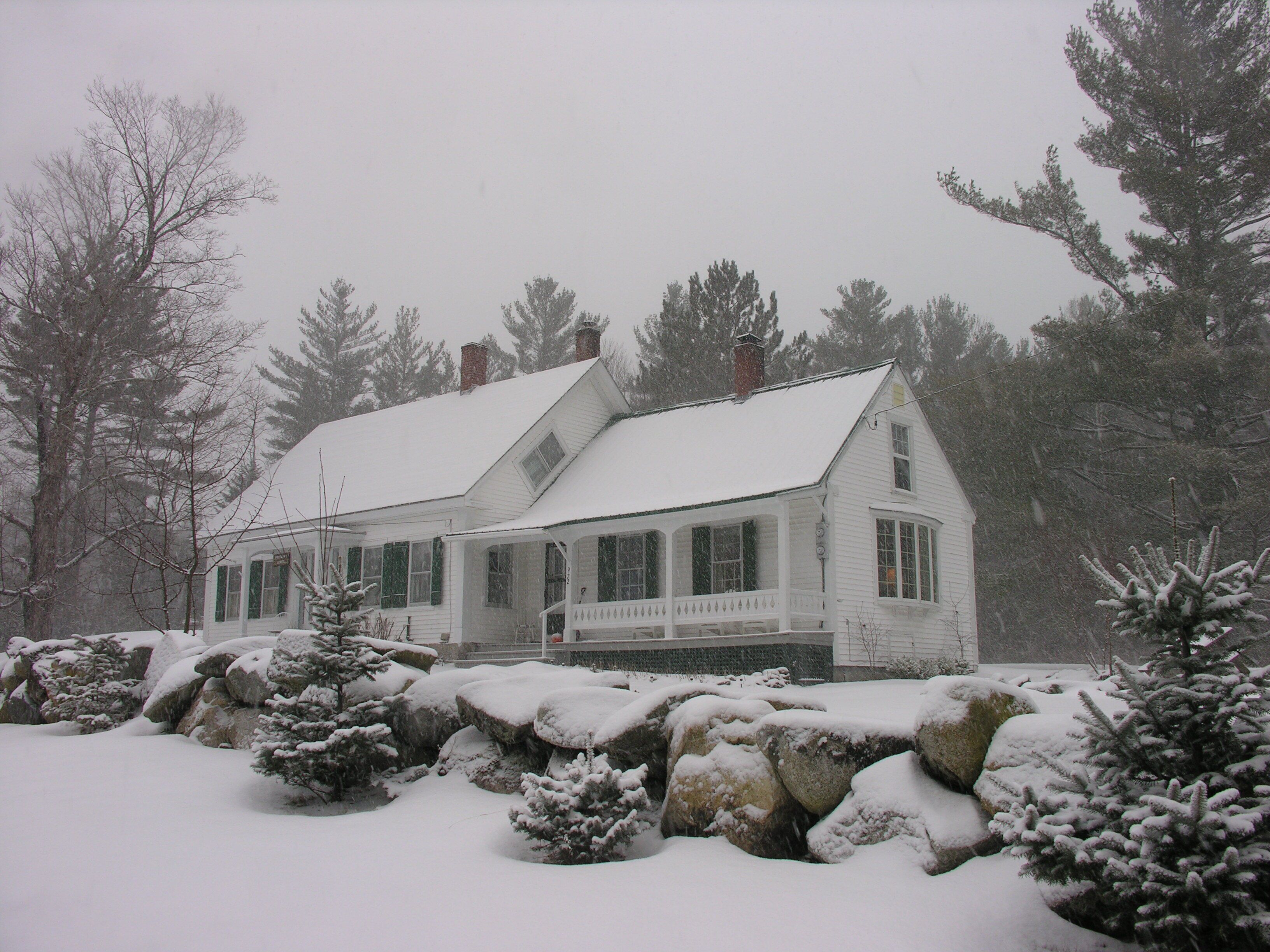 A white house with a snow-covered roof and porch, surrounded by snow-covered rocks and evergreen trees.