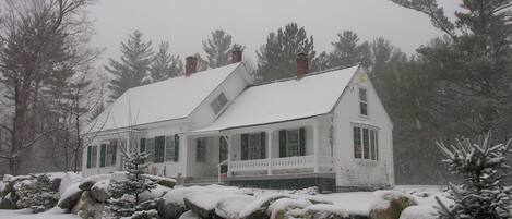 A white house with a snow-covered roof and porch, surrounded by snow-covered rocks and evergreen trees.