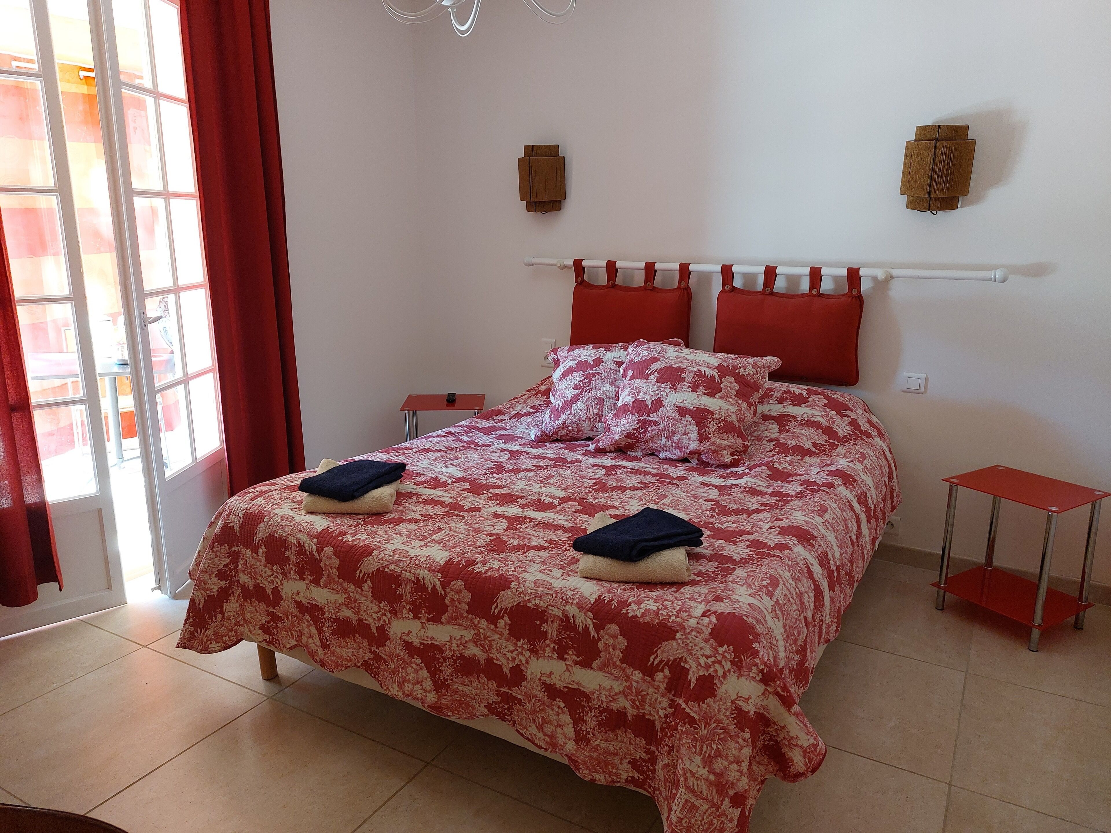 Une chambre à coucher avec un lit, des rideaux rouges, une petite table et une étagère fixée au mur.