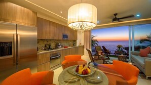 Una cocina moderna con una isla grande, un comedor con sillas color naranja y vistas al océano al atardecer.