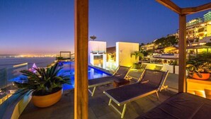 Zona de piscina en la azotea con sillas de descanso y vistas a la ciudad al anochecer.