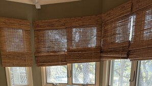 Een kamer met een bruine leren bank, een ronde houten tafel en meerdere ramen met bamboe blinds.