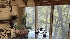 Een eethoek met een glazen tafel, omringd door stoelen, een decoratieve lamp en uitzicht op bomen door het raam.