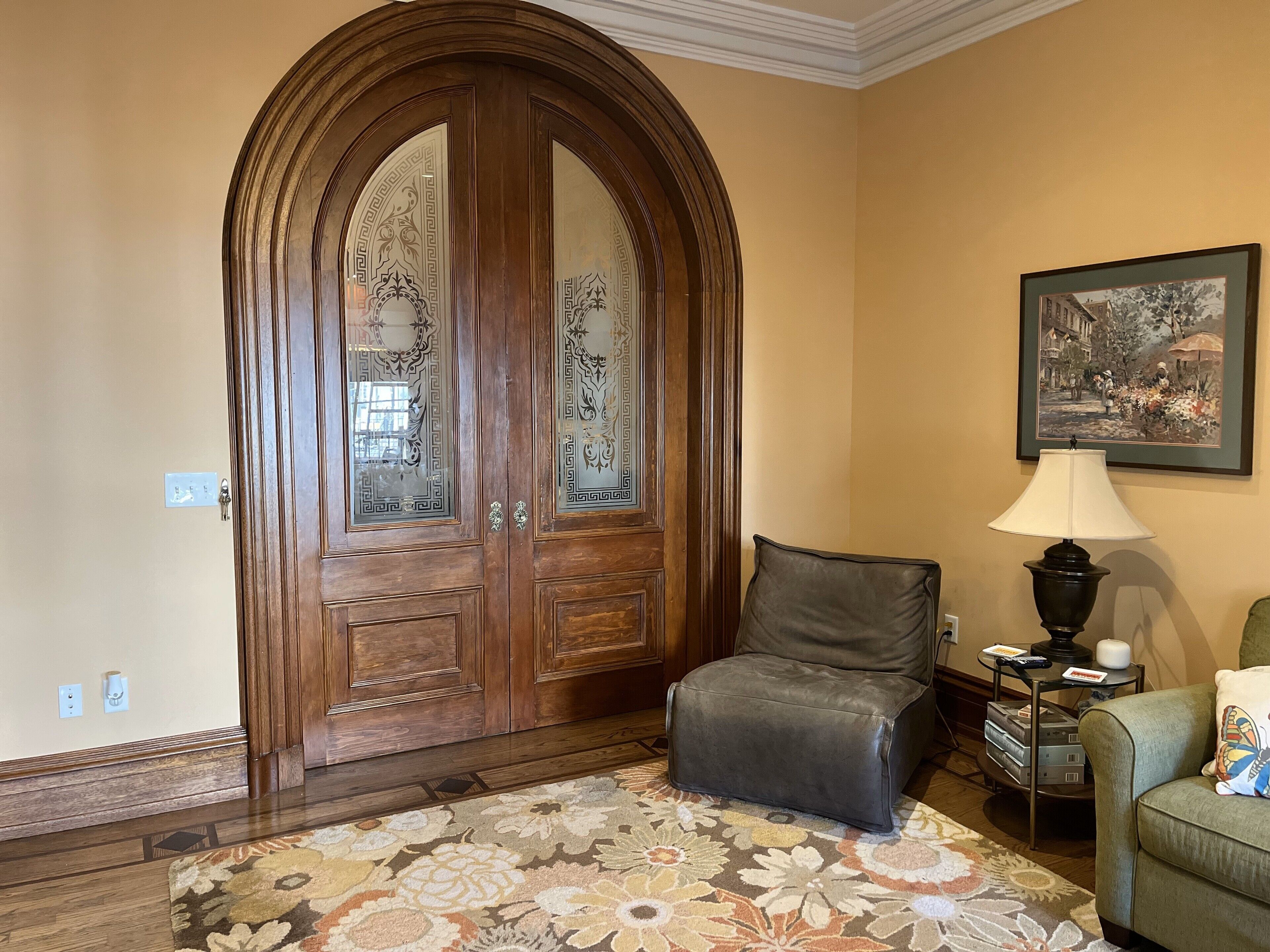 A room with a large wooden door with glass panels, a leather chair, a patterned rug, a lamp, and a framed picture on the wall.