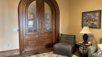 A room with a large wooden door with glass panels, a leather chair, a patterned rug, a lamp, and a framed picture on the wall.