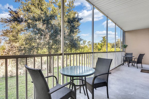 A covered outdoor area with a round glass table and two chairs, a small table with chairs, and a view of trees and sky.