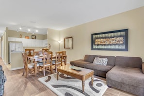A living room with a brown sofa, wooden dining table, and chairs. There is a kitchen in the background with white cabinets and a refrigerator.