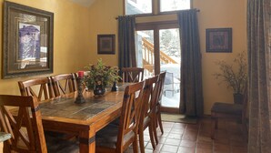 Un comedor con mesa de madera, sillas y un candelabro. Hay dos ventanas con cortinas y una puerta que da al exterior.