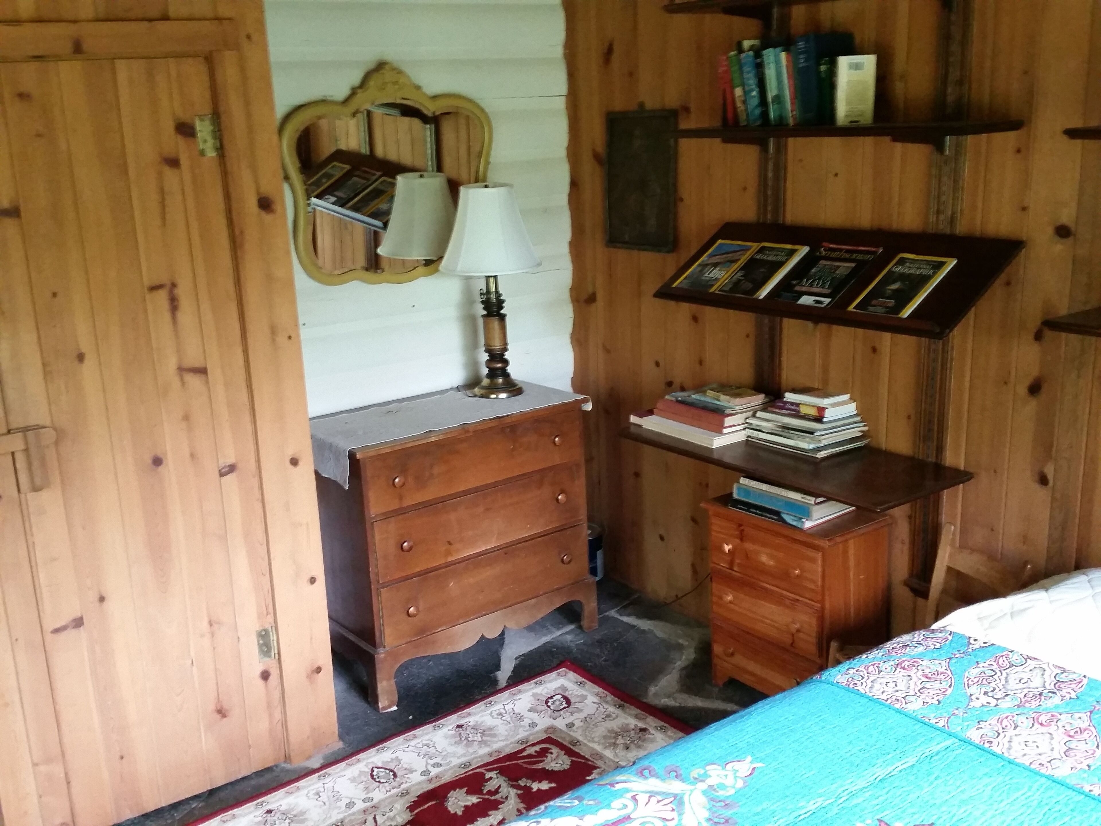 A wooden room with a bed, a dresser, a mirror, and a shelf with books.