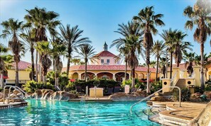 A pool area with clear blue water, palm trees, and a building with a red roof.