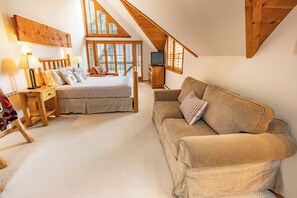 A cozy bedroom with a bed, a wooden headboard, a nightstand, a bedside table with lamps, a beige sofa, and a TV on a wooden cabinet. - Pinnacle Ridge, 3 Bedroom + Den (Whistler)