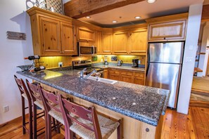 A kitchen with wooden cabinets, granite countertops, stainless steel appliances, and bar stools. - Pinnacle Ridge, 3 Bedroom + Den (Whistler)
